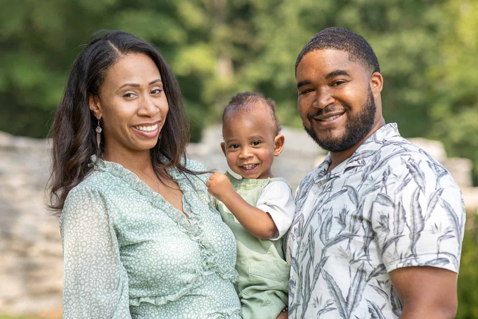 Black couple smiling and holding their one year old baby at the park.